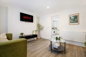 Una sala de estar moderna con una televisión de pantalla plana mostrando 'NETFLIX', una mesa de centro de vidrio con libros, un sillón verde, una planta en maceta y un cuadro en la pared.