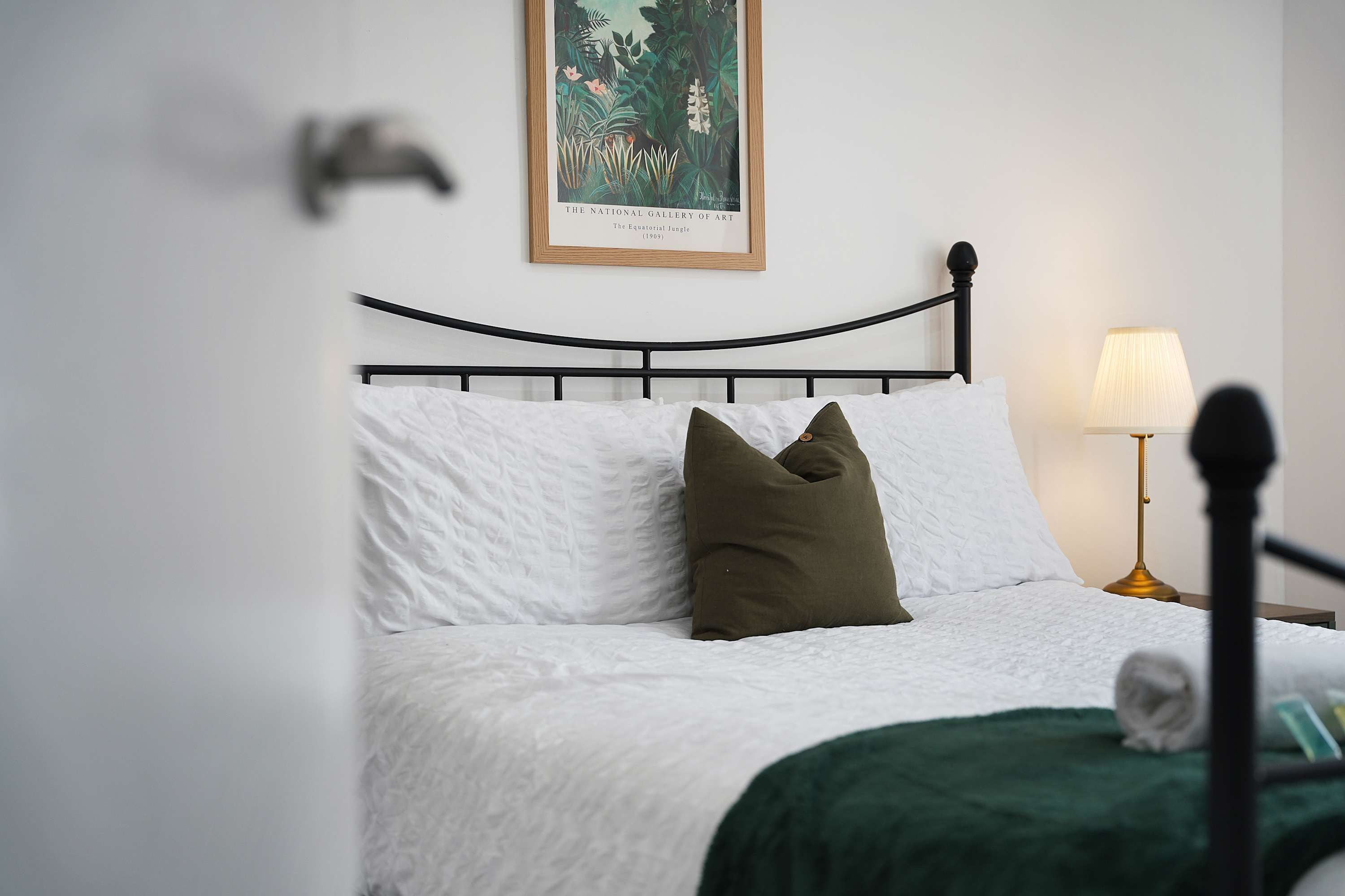 A neatly made bed with white bedding and a green throw blanket, a black metal headboard, and a framed painting above the bed.