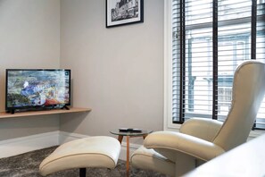 A living room with a television on a stand, a chair, a small table, and a window with blinds. - Luxury 1 Bedroom in Hartlepool With Wifi (Hartlepool)