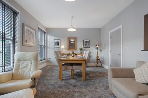 A dining area with a table set for four, surrounded by chairs, with a lamp and framed pictures on the walls. - Luxury 1 Bedroom in Hartlepool With Wifi (Hartlepool)