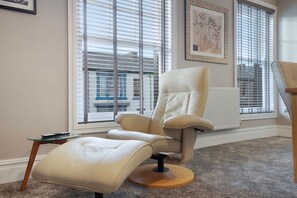 Una habitación con un ventanal grande, un sillón blanco, una mesita y un cuadro enmarcado en la pared.