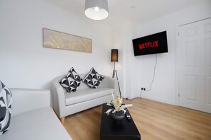 A modern living room with a white sofa, a black coffee table with a vase, a floor lamp, and a wall-mounted TV displaying 'NETFLIX'. - Spacious 4BR Home Sleeps 8 by Stay With Us (Consett)