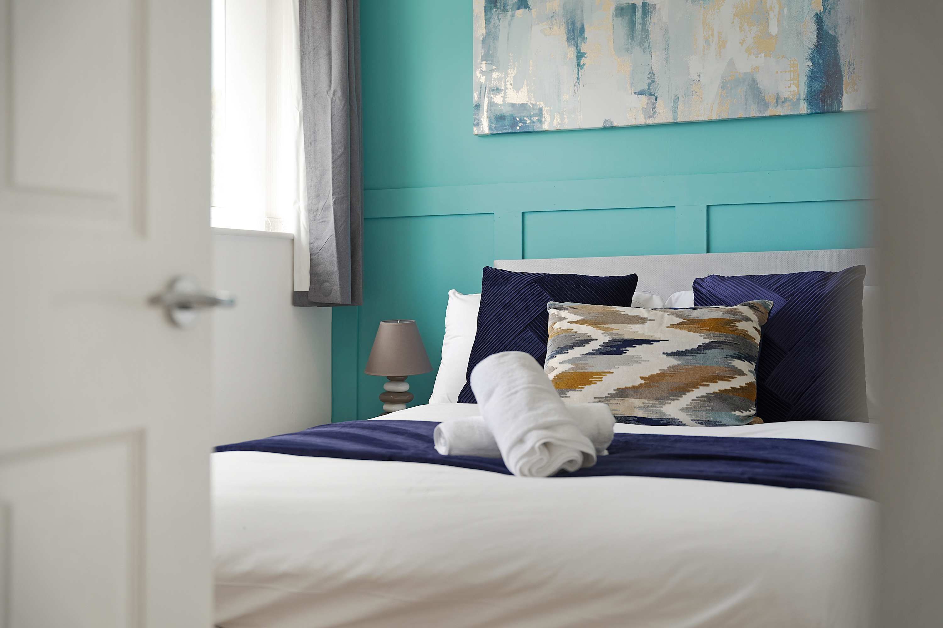 Une chambre d’hôtel avec un lit, une fenêtre avec des rideaux, un mur orné d’un tableau et une table de chevet avec une lampe.