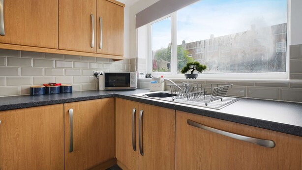 A modern kitchen with wooden cabinets, a microwave, a sink, and a window with a view of the city. - Spacious 4BR Home Sleeps 8 by Stay With Us (Consett)