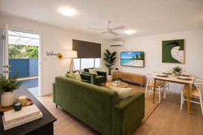 Ein Wohnzimmer mit einem grünen Sofa, einem Esstisch für zwei Personen, einem Deckenventilator und einem Fernseher an der Wand.