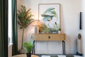 Una habitación con una consola de madera, una obra de arte enmarcada de un pájaro, una lámpara y plantas en macetas.
