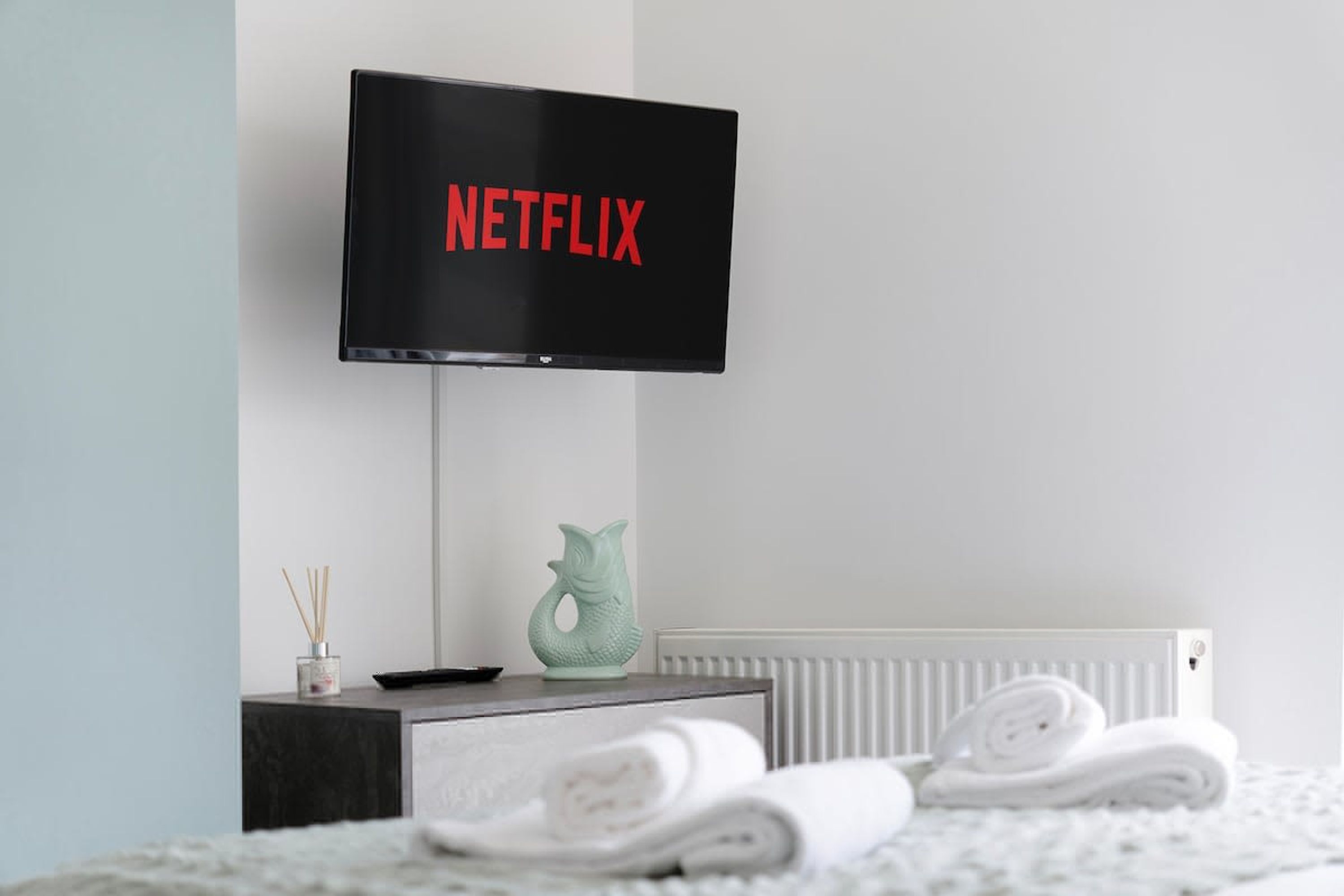 A modern room with a flat-screen TV displaying 'NETFLIX' on the screen, a small table with a decorative fish sculpture, and neatly folded towels on the floor.