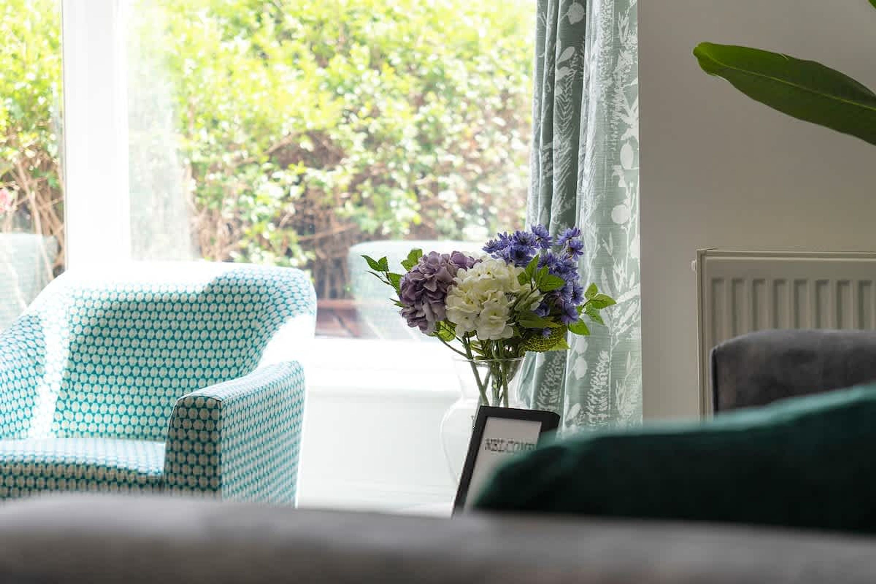 A room with a patterned armchair, a vase with flowers, and a view of greenery outside.