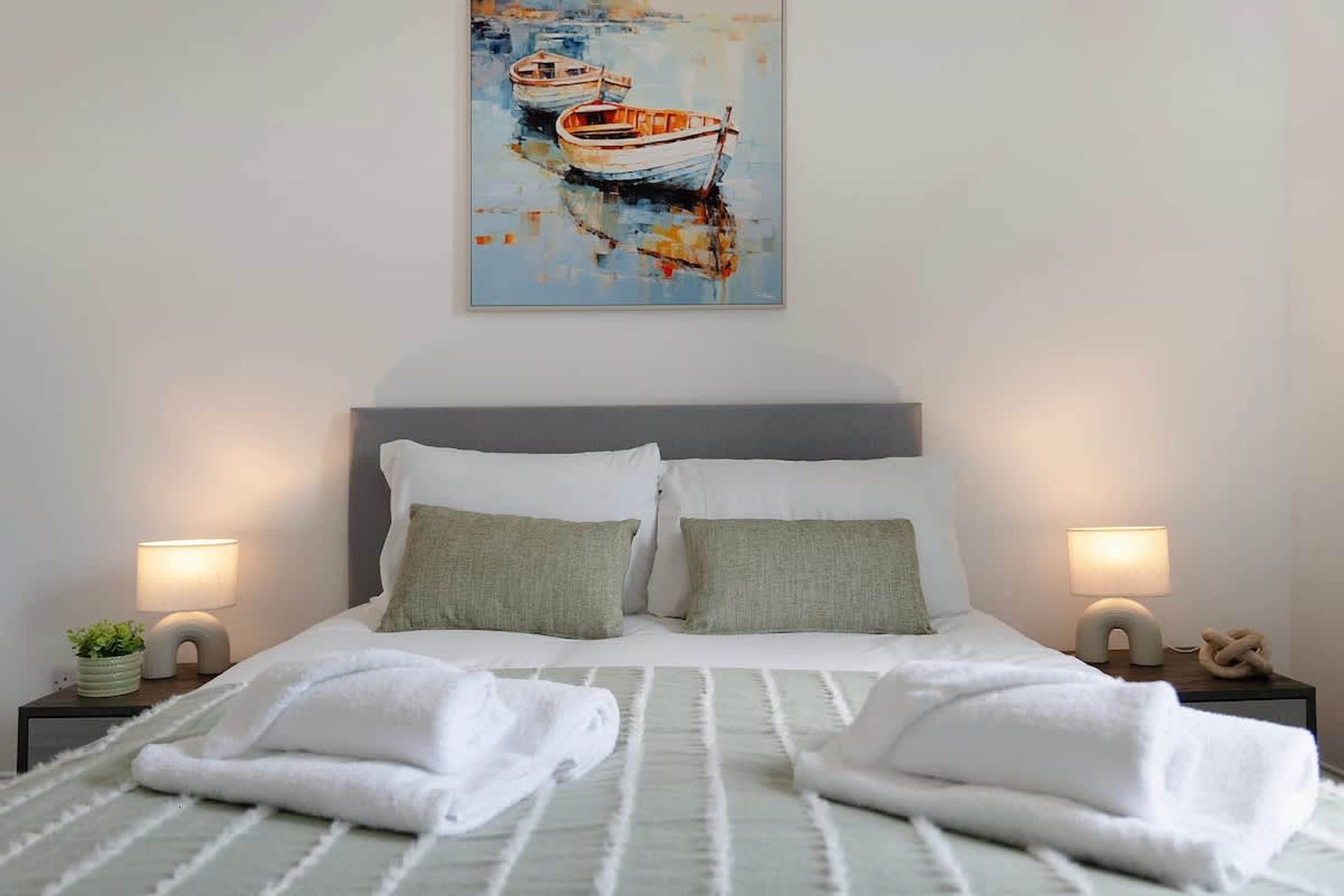Una cama bien hecha con almohadas, dos lámparas de noche y un cuadro de barcos en la pared.