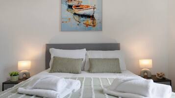 Una cama bien hecha con almohadas, dos lámparas de noche y un cuadro de barcos en la pared.