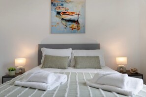 Una cama bien hecha con almohadas, dos lámparas de noche y un cuadro de barcos en la pared.