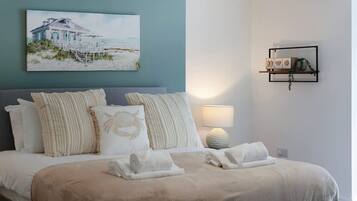 Una habitación de hotel con una cama, un cuadro de una casa de playa, un estante montado en la pared con adornos y una lámpara de noche.