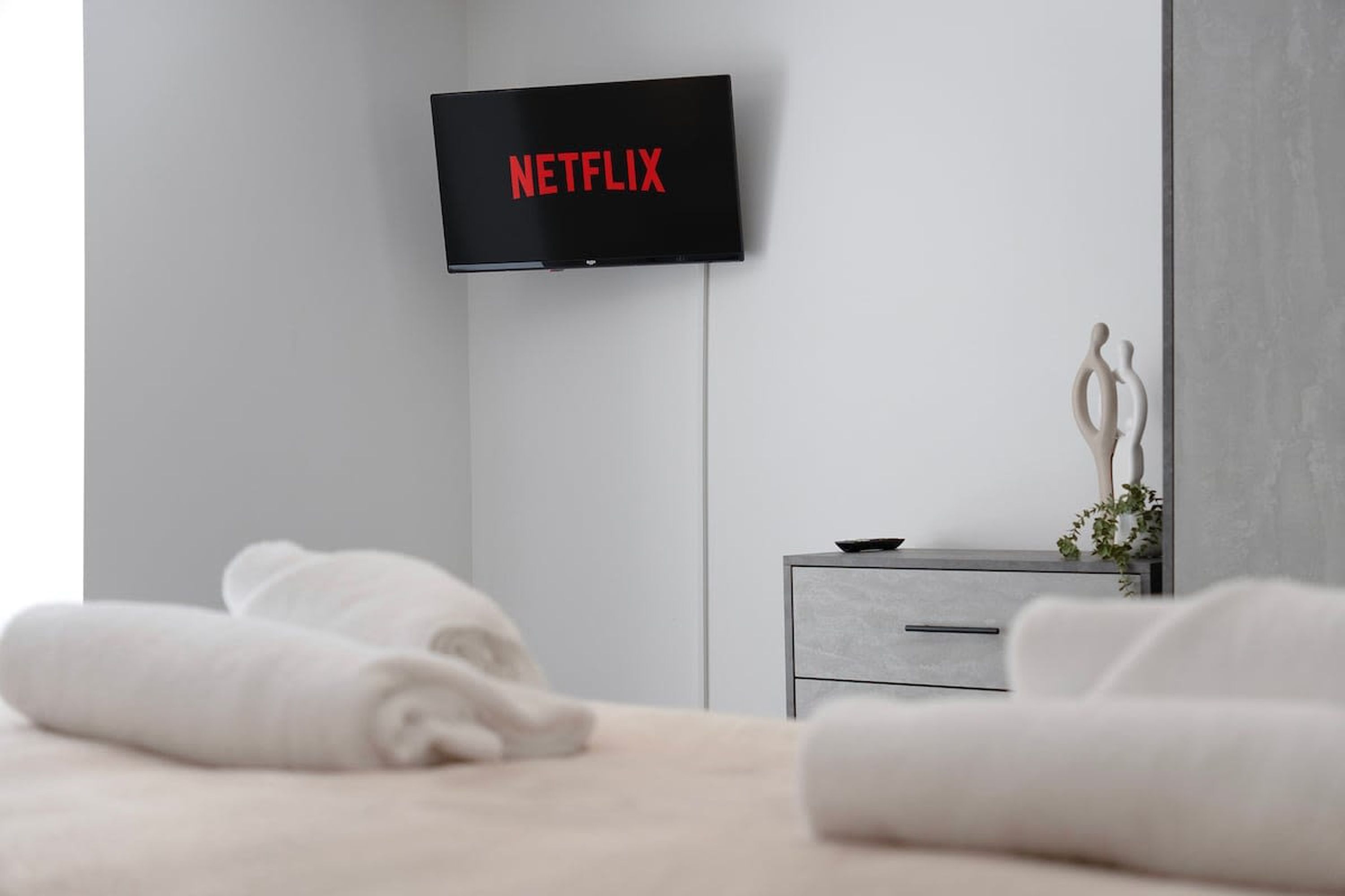 Habitación de hotel moderna con una televisión de pantalla plana montada en la pared mostrando el logo de Netflix, una mesita de noche gris con una planta y toallas blancas dobladas con cuidado sobre la cama.