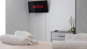 Habitación de hotel moderna con una televisión de pantalla plana montada en la pared mostrando el logo de Netflix, una mesita de noche gris con una planta y toallas blancas dobladas con cuidado sobre la cama.