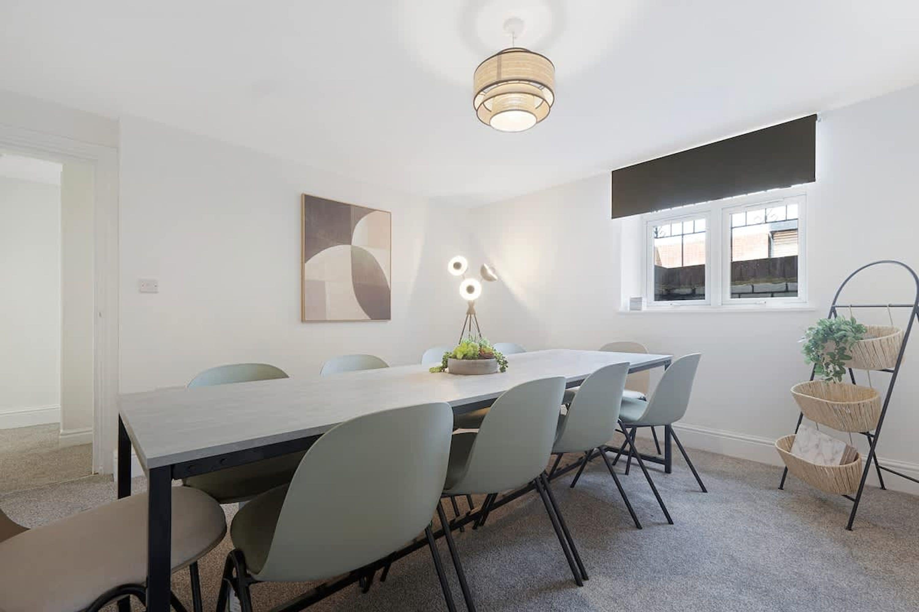 A modern dining room with a long table, six chairs, a hanging light fixture, and a window with blinds.