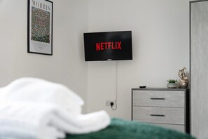 Un dormitorio con cama, cómoda, televisor de pared mostrando 'NETFLIX' y un cuadro en la pared.