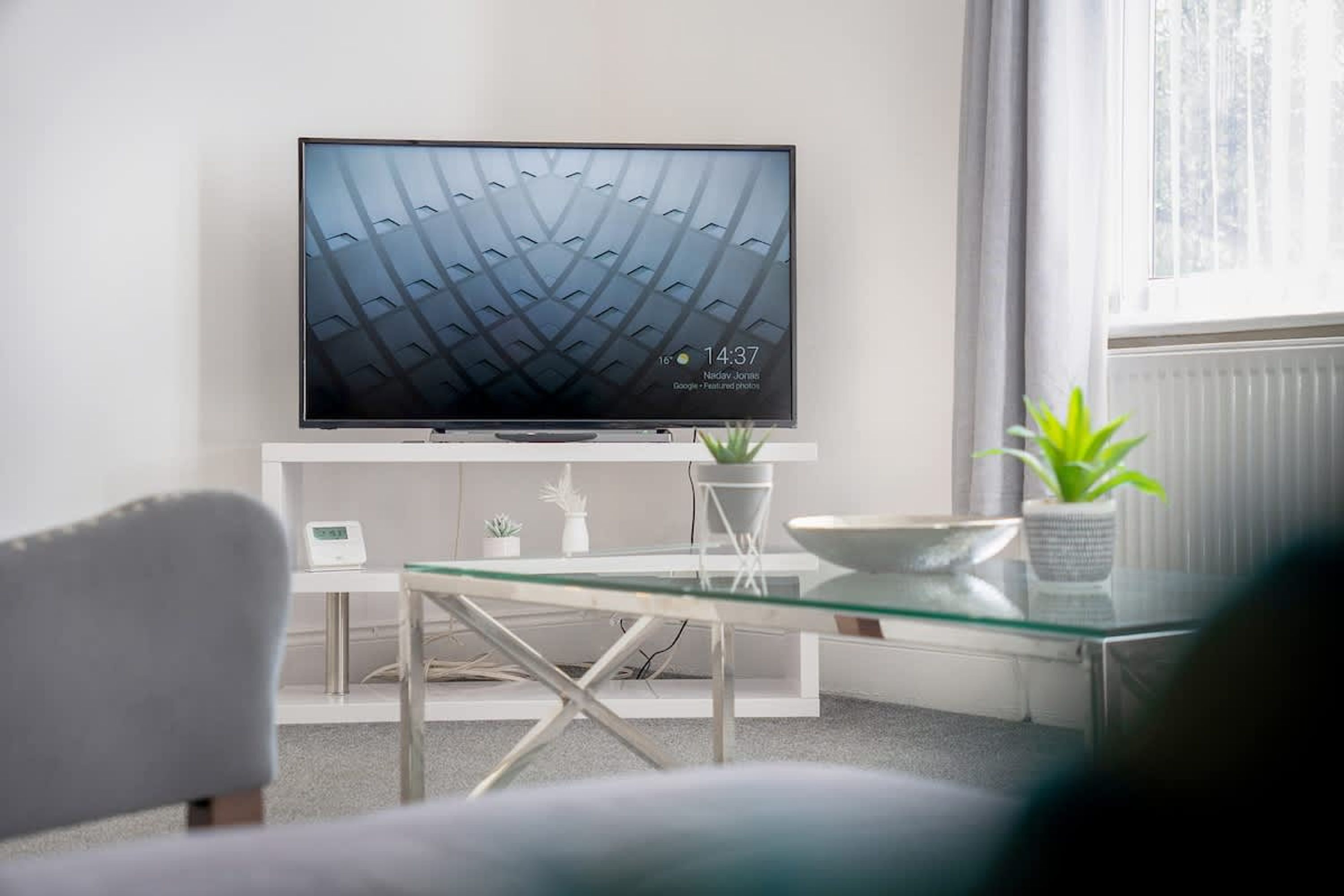 Una sala de estar moderna con un televisor de pantalla plana sobre un mueble blanco, una mesa de centro de vidrio, un sofá blanco y una planta en maceta.