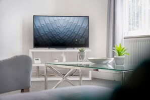 Ein modernes Wohnzimmer mit einem Flachbildfernseher auf einem weißen Standfuß, einem Glastisch, einem weißen Sofa und einer Topfpflanze.