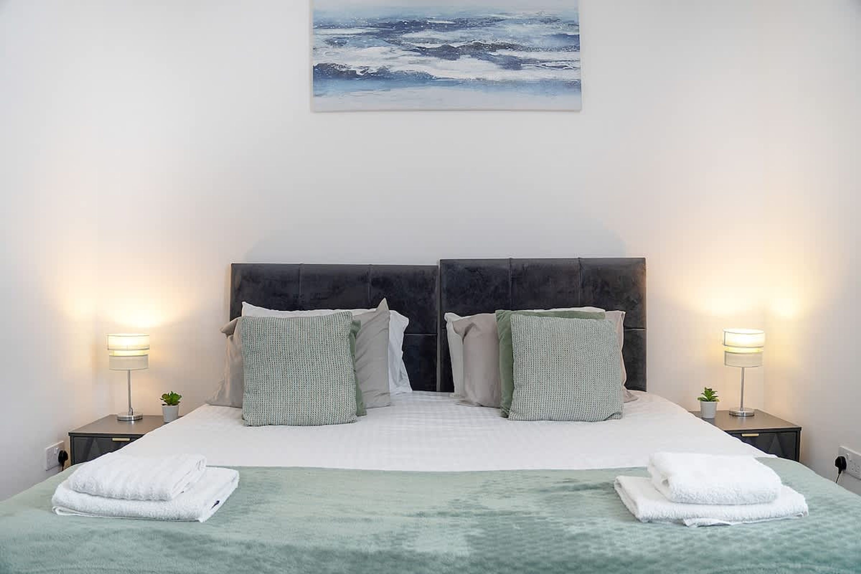 A bed with a green blanket, two pillows, two bedside lamps, and a painting above.