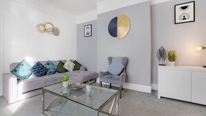Un salon moderne avec un canapé gris, une table basse en verre et un fauteuil gris. On y trouve des coussins décoratifs, une œuvre d’art circulaire accrochée au mur et un tableau encadré.