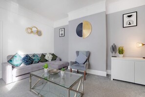 A modern living room with a grey sofa, a glass coffee table, and a grey armchair. There are decorative pillows, a wall-mounted circular artwork, and a framed picture on the wall. - Family Three Bedroom Home Five Min Walk to Metro (South Shields)