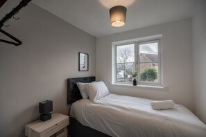 Ein Schlafzimmer mit einem Bett, einem Nachttisch mit Lampe, einem Fenster mit Blick auf Häuser und einem gerahmten Bild an der Wand.