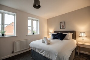 A hotel room with a bed, two bedside lamps, a radiator, and windows. - Brecon Gardens Horizon Stays (Middlesbrough)