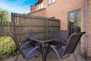 A patio with a glass table and four chairs, a wooden fence, and a brick building with a door. - Modern 3BR Brompton Court Wi-fi By Stay With Us (Richmond)