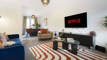 Una sala de estar moderna con un sofá, una mesa de centro, un televisor montado en la pared y un comedor al fondo.