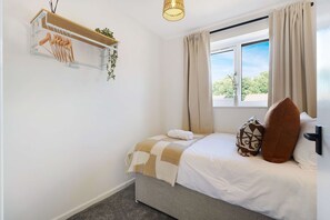 A bedroom with a bed, a hanging plant, a window with curtains, and a wall-mounted shelf with clothes. - Modern 3BR Brompton Court Wi-fi By Stay With Us (Richmond)
