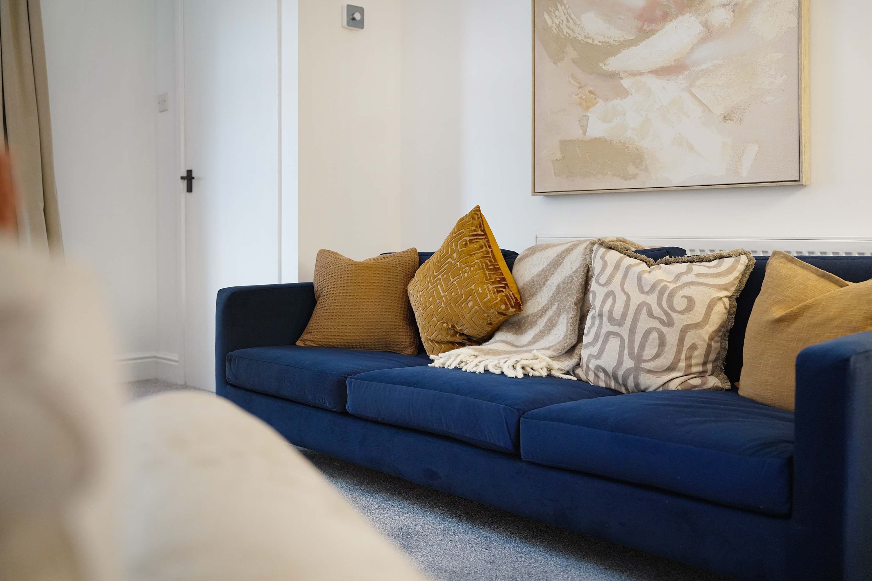A blue sofa with patterned cushions, a white wall, and a framed abstract painting.