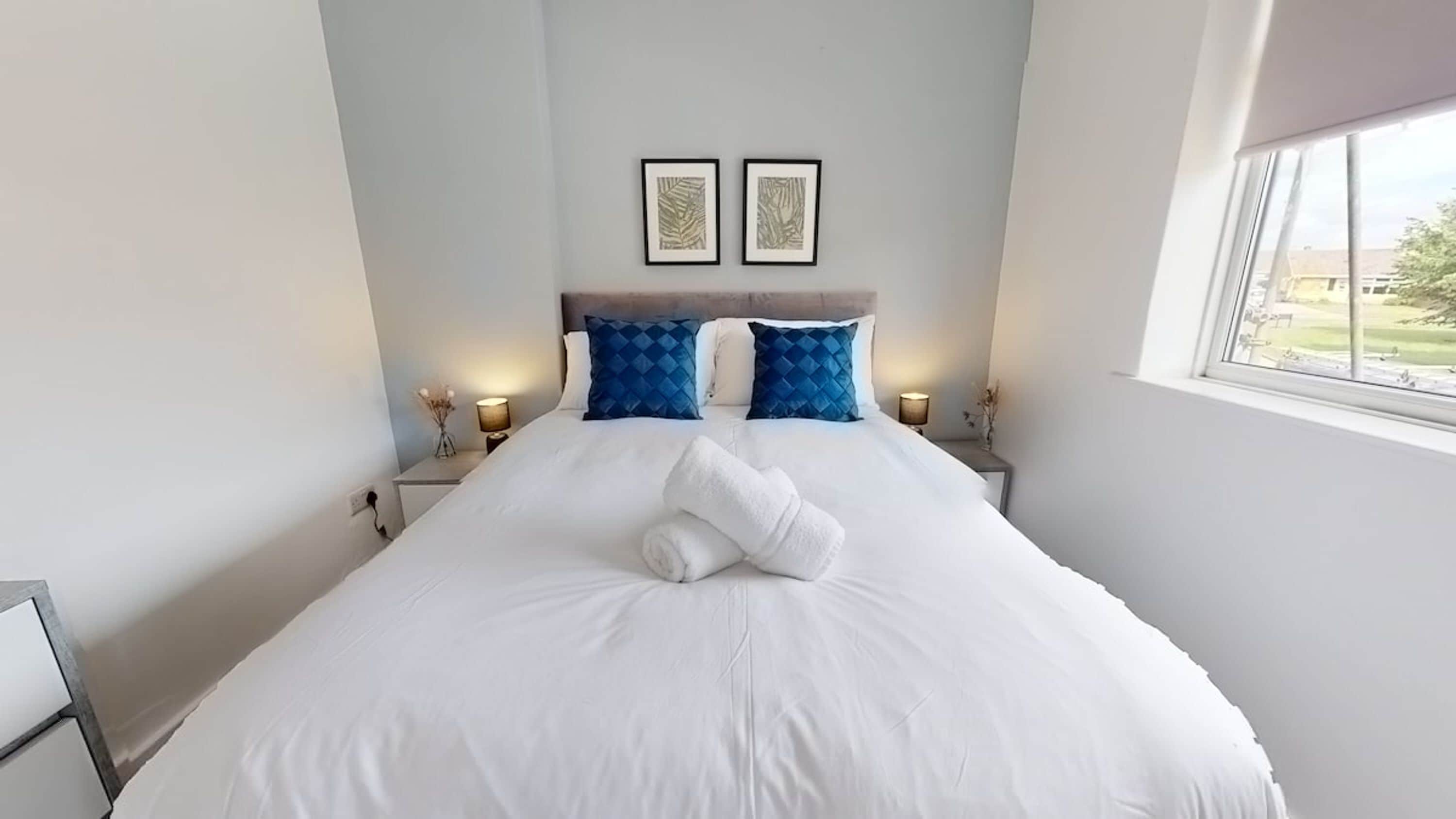 A neatly made bed with white linens and two blue pillows, a nightstand with a lamp, and two framed artworks on the wall.