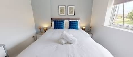 A neatly made bed with white linens and two blue pillows, a nightstand with a lamp, and two framed artworks on the wall.