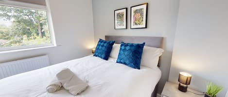 A neatly made bed with a white duvet and two blue pillows, a bedside table with a lamp, and a framed picture on the wall.