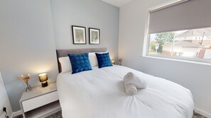 A hotel room with a bed, two pillows, a bedside table with a lamp, and a window with a view of the outside. - Daventry House Horizon Stays (Stockton-on-Tees)
