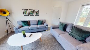Ein Wohnzimmer mit einem grauen Sofa, einem weißen Couchtisch und einer Stehlampe. An der Wand hängen drei gerahmte Bilder.