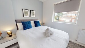 A neatly made bed with white linens and blue pillows, a bedside table with a lamp, and a window with a view of buildings. - Daventry House Horizon Stays (Stockton-on-Tees)