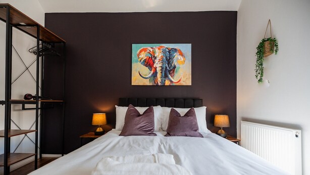 A hotel room with a large bed, two bedside lamps, a painting of an elephant, a radiator, and a hanging plant. - The Cockton Quarters Horizon Stays (Bishop Auckland)