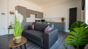 A modern living room with a grey sofa, a small wooden table with a plant, and a kitchen area with grey cabinets and white tiles. - The Cockton Quarters Horizon Stays (Bishop Auckland)