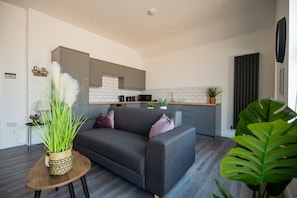 Ein modernes Wohnzimmer mit einem grauen Sofa, einem kleinen Holztisch mit einer Pflanze und einer Küchenzeile mit grauen Schränken und weißen Fliesen.