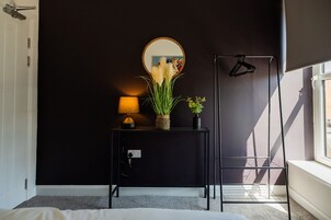 Un dormitorio con pared negra, una mesita de noche con lámpara, un espejo y una planta en maceta.