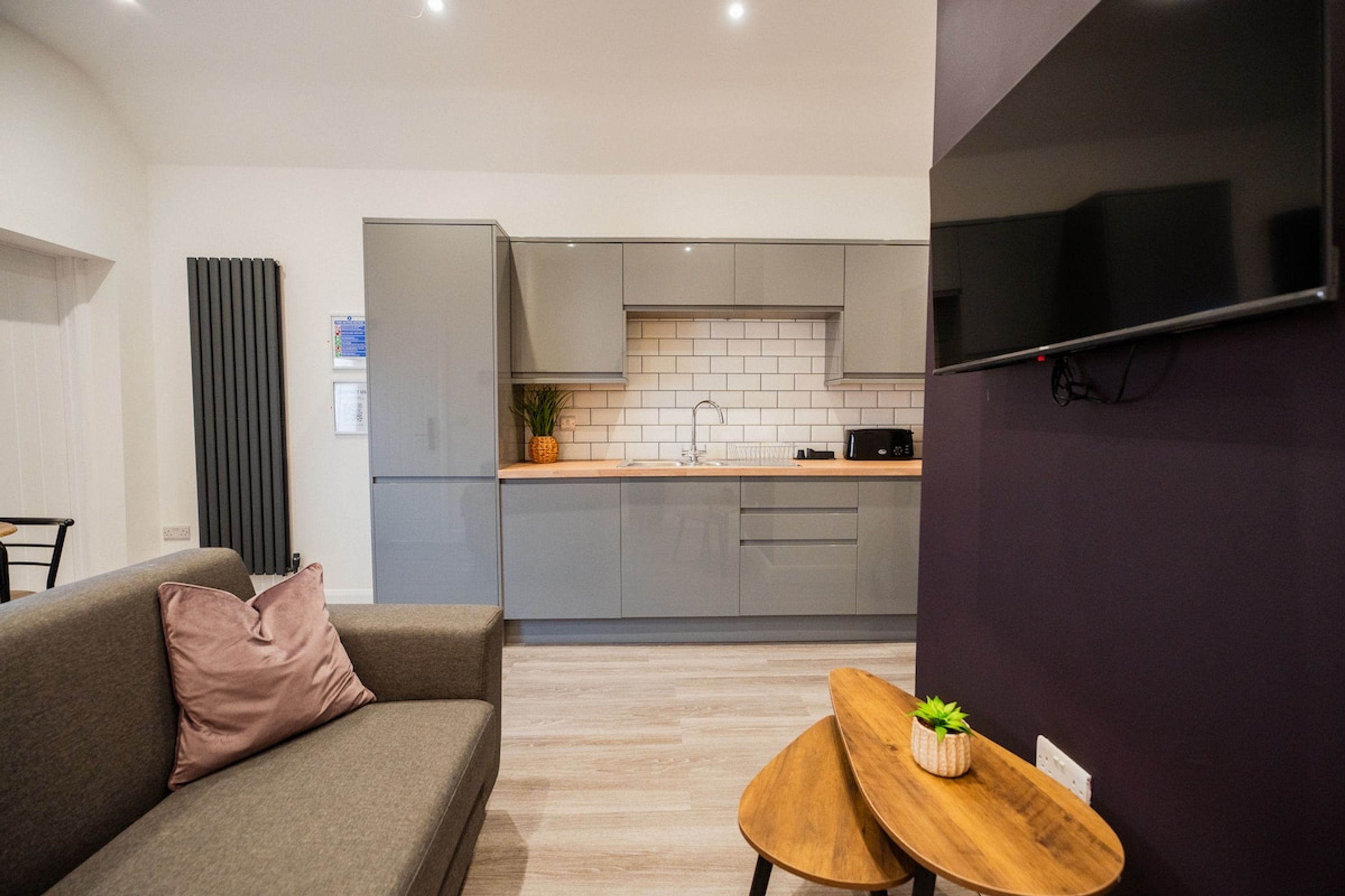 Un salon moderne avec un canapé et une petite table, une cuisine avec des meubles gris et un dosseret carrelé, et un téléviseur à écran plat fixé sur un mur foncé.