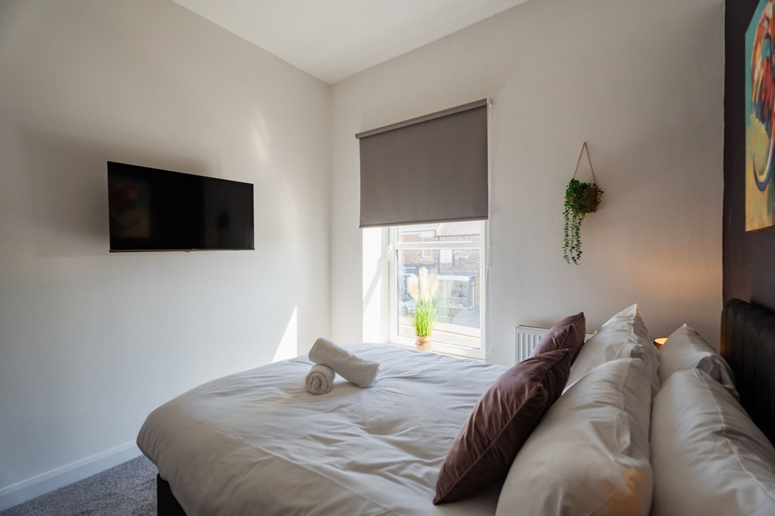 A bedroom with a bed, a television mounted on the wall, a window with a view of buildings, and a hanging plant.