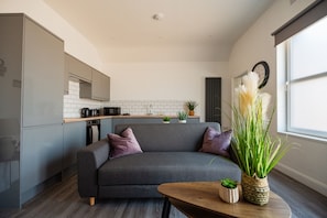 Um espaço de estar moderno com sofá cinzento, mesa de centro de madeira e cozinha com azulejos brancos e exaustor preto.