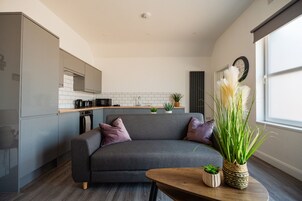 Un espacio moderno para vivir con un sofá gris, una mesa de centro de madera y una zona de cocina con azulejos blancos y una campana extractora negra.