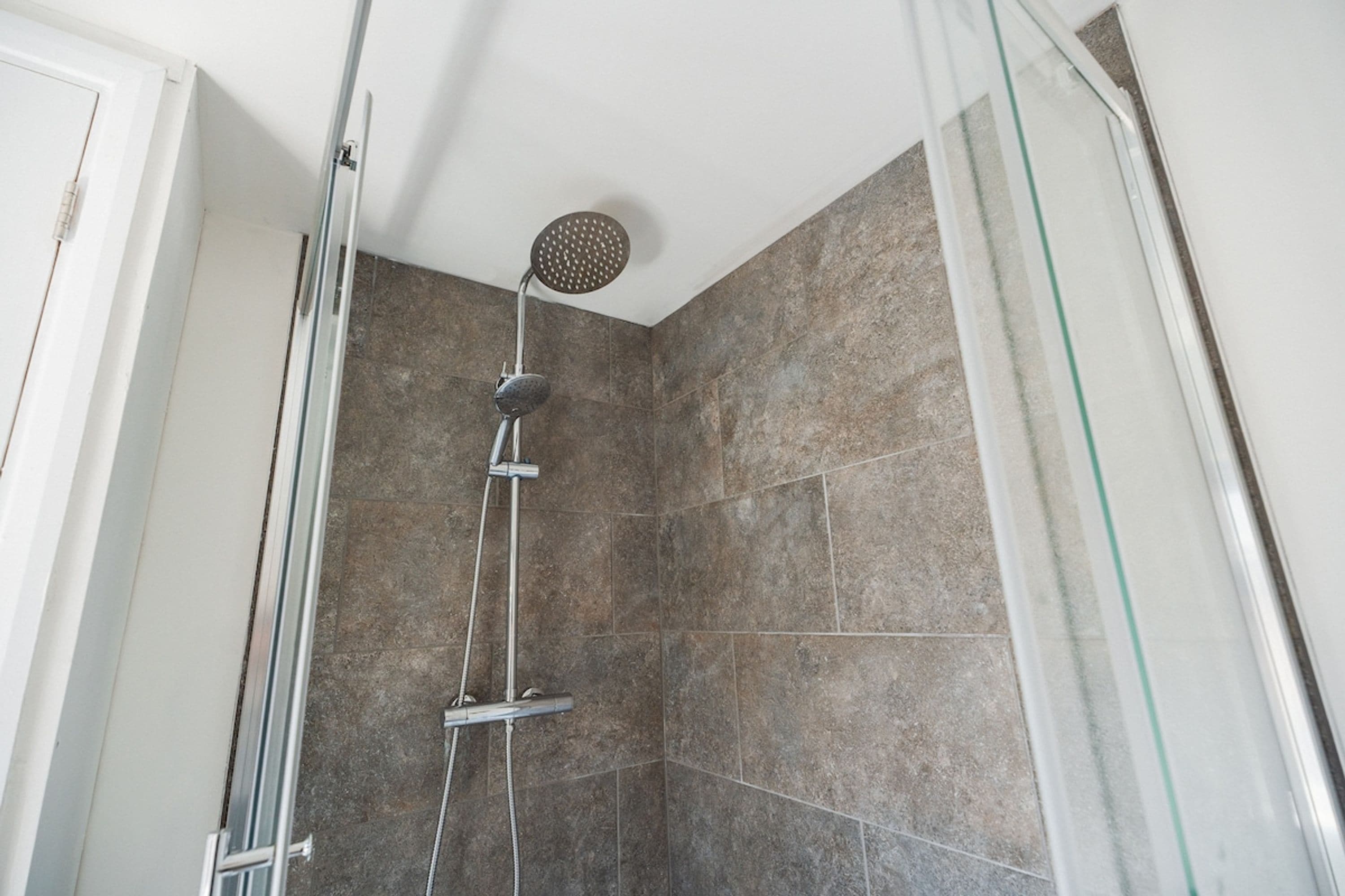 A shower with a handheld showerhead and a rain shower head, set in a tiled room with a glass door.