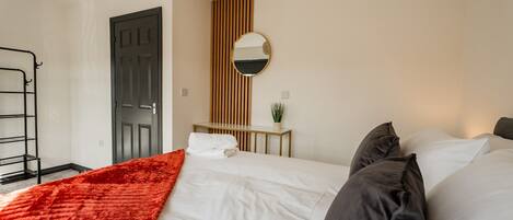 Habitación de hotel con una cama, una mesita de noche, un espejo, una puerta y un estante fijado a la pared.