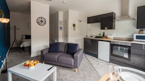 A modern apartment with a grey sofa, a dining table with food, a kitchen with appliances, and a clock on the wall. - 1 Bedroom Studio Apartment Central Middlesbrough (Middlesbrough)