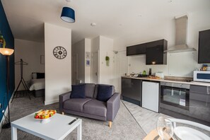 Ein modernes Apartment mit grauem Sofa, einem Esstisch mit Speisen, einer Küche mit Geräten und einer Uhr an der Wand.