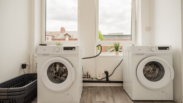 A laundry room with two front-loading washing machines, a black laundry basket, and a window with a view of buildings. - 1 Bedroom Studio Apartment Central Middlesbrough (Middlesbrough)